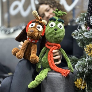 Puede incluir: Dos juguetes de peluche tejidos a crochet, un reno marrón y un Grinch verde, están sentados en un sofá gris. El reno tiene una nariz roja y una bufanda roja y blanca. El Grinch tiene una bufanda roja y un sombrero verde.