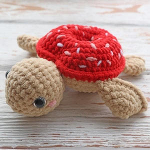 May include: A crocheted turtle toy with a red and white donut shell. The turtle has a brown body with black eyes and a pink nose.