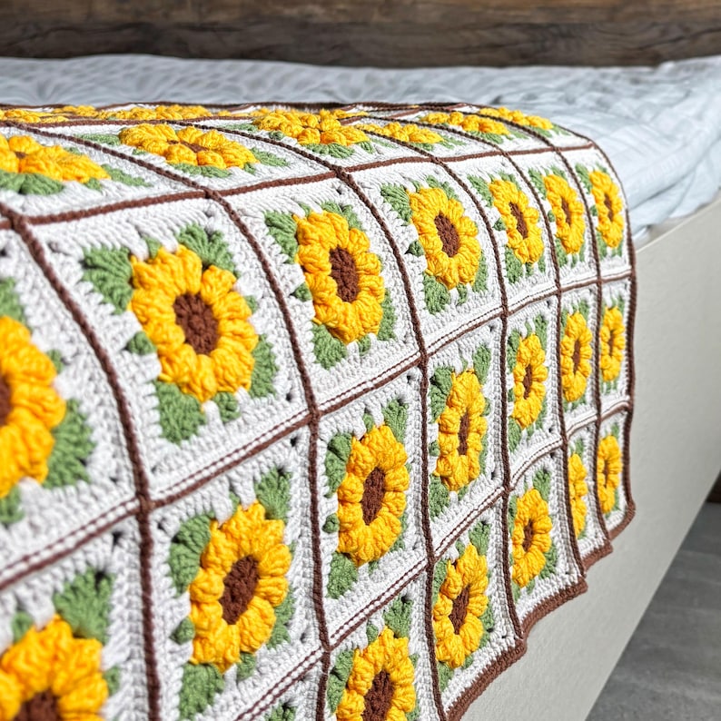 K&ouml;nnte beinhalten: Eine handgefertigte H&auml;keldecke mit einem sich wiederholenden Sonnenblumenmuster. Die Decke hat einen wei&szlig;en Hintergrund mit gelben Sonnenblumen, gr&uuml;nen Bl&auml;ttern und braunen R&auml;ndern. Die Decke ist &uuml;ber ein Bett gelegt.