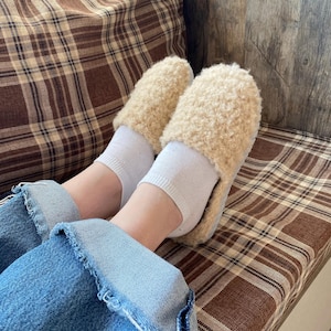 May include: A pair of fluffy beige slippers with a soft, fuzzy texture. The slippers are worn by a person with white socks and blue jeans.