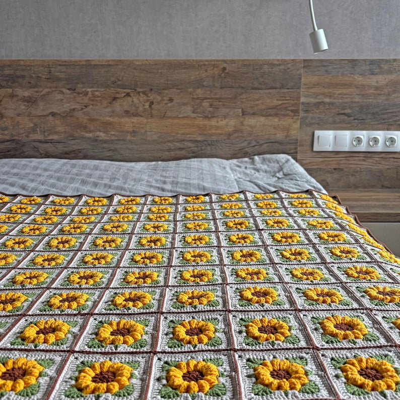 K&ouml;nnte beinhalten: Eine handgefertigte H&auml;keldecke mit einem sich wiederholenden Sonnenblumenmuster. Die Decke besteht aus gelber, brauner, gr&uuml;ner und wei&szlig;er Wolle. Die Decke liegt auf einem Bett mit einem h&ouml;lzernen Kopfteil.