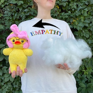 May include: A yellow crocheted duck wearing glasses and a pink bow. The duck is held in front of a person wearing a white sweatshirt with the word "EMPATHY" in rainbow letters. A cloud of white stuffing is held in the other hand.