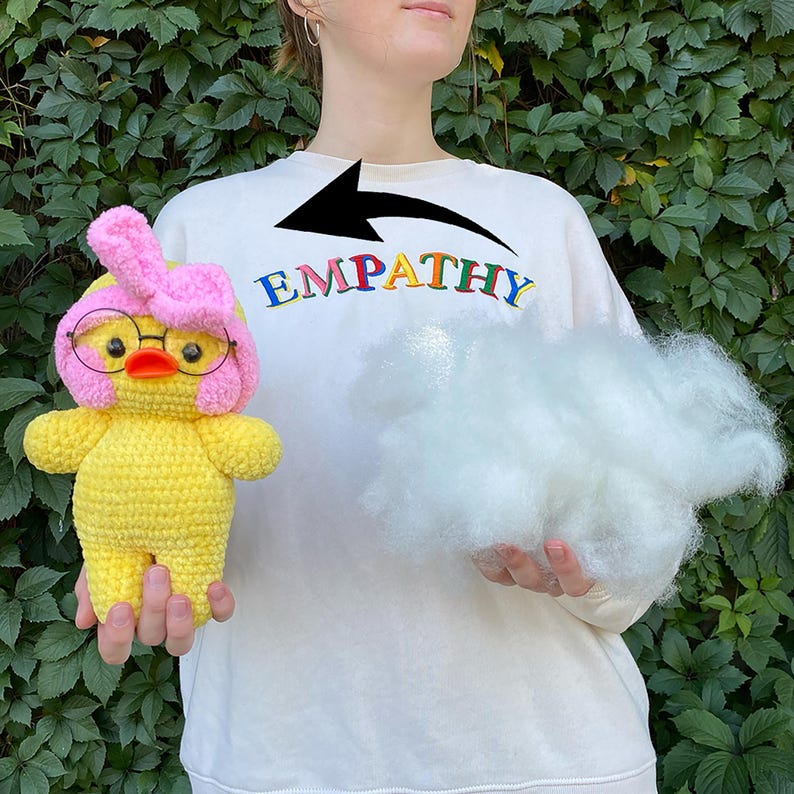 May include: A yellow crocheted duck wearing glasses and a pink bow. The duck is held in front of a person wearing a white sweatshirt with the word "EMPATHY" in rainbow letters. A cloud of white stuffing is held in the other hand.