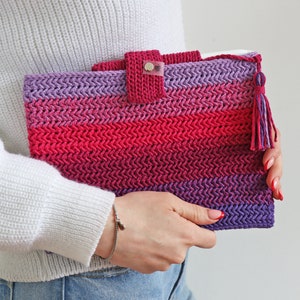 Pode incluir: Uma capa de tablet de crochê em um gradiente de tons rosa, vermelho e roxo. A capa apresenta um padrão de ponto texturizado, um fecho de aba com botão e uma borla combinando. A capa é segurada por uma pessoa vestindo um suéter branco.