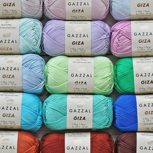 May include: A collection of 12 skeins of Gazzal Giza yarn in various colors, including light blue, lavender, green, turquoise, and blue. Each skein is labeled with the brand name, yarn name, weight, and yardage. The yarn is 100% mercerized cotton.