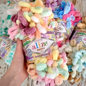 May include: A close-up of Alize Puffy Color yarn in a variety of pastel colors, including pink, yellow, green, and orange. The yarn is displayed in a cylindrical container with the brand name and product details. The image showcases the soft, textured loops of the yarn.