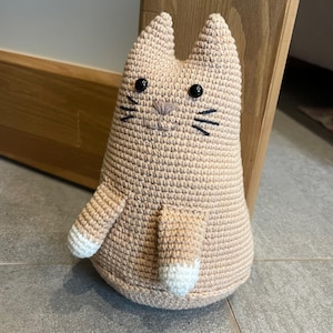 Op de afbeelding: Een handgemaakte, licht perzik kleurige gehaakte kattenfiguur. De kat heeft zwarte ogen en snorharen, met witte accenten op de poten en staart. Een grillig, decoratief item.