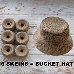 K&ouml;nnte beinhalten: Sechs Kn&auml;uel braunes Garn sind in zwei Reihen zu je drei angeordnet. Neben dem Garn befindet sich ein brauner geh&auml;kelter Bucket-Hut. Der Text "6 SKEINS = BUCKET HAT" ist unter dem Bild gedruckt.