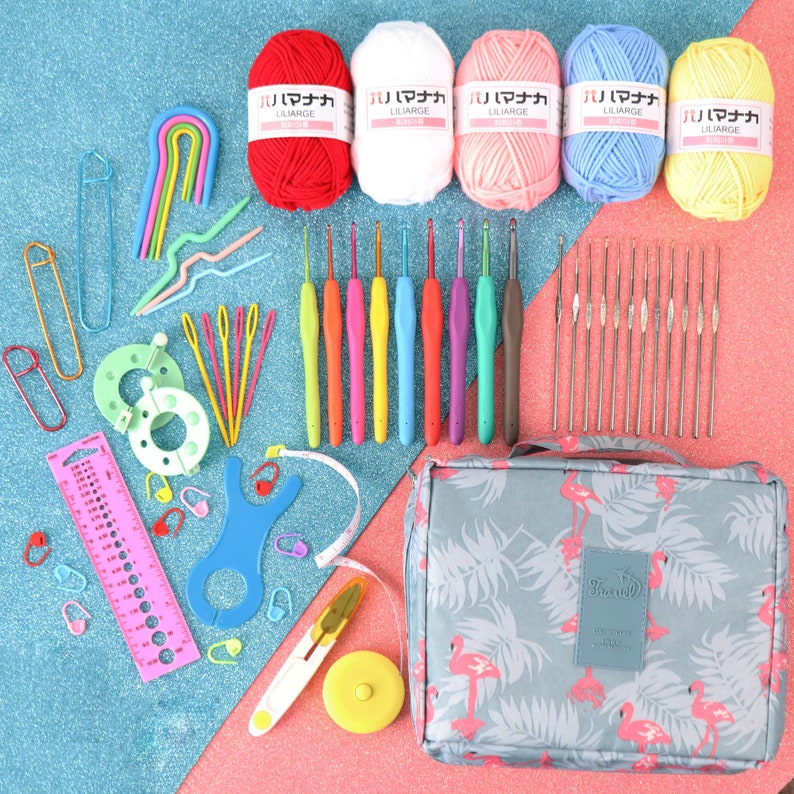 May include: A set of colourful crochet hooks, yarn, and other crochet supplies are arranged on a blue and pink glittery surface. The supplies are organised in a flamingo-patterned zippered pouch. The yarn is in red, white, pink, blue, and yellow. The crochet hooks are in various colours, including green, blue, orange, yellow, and purple. The pouch is grey with pink flamingos and green leaves.