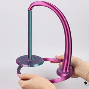 Op de afbeelding: Een 3D-geprinte, iriserende wijnglashouder. De houder heeft een verticale steun die overloopt van teal naar paars, verbonden met een gebogen boog en een cirkelvormige basis. De boog en de basis hebben een verloop van paars en roze.