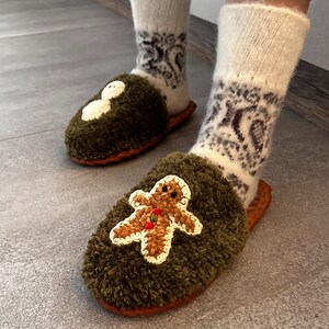 May include: A pair of green fuzzy slippers with a snowman and gingerbread man design. The slippers are worn by a person wearing white socks with a brown pattern.