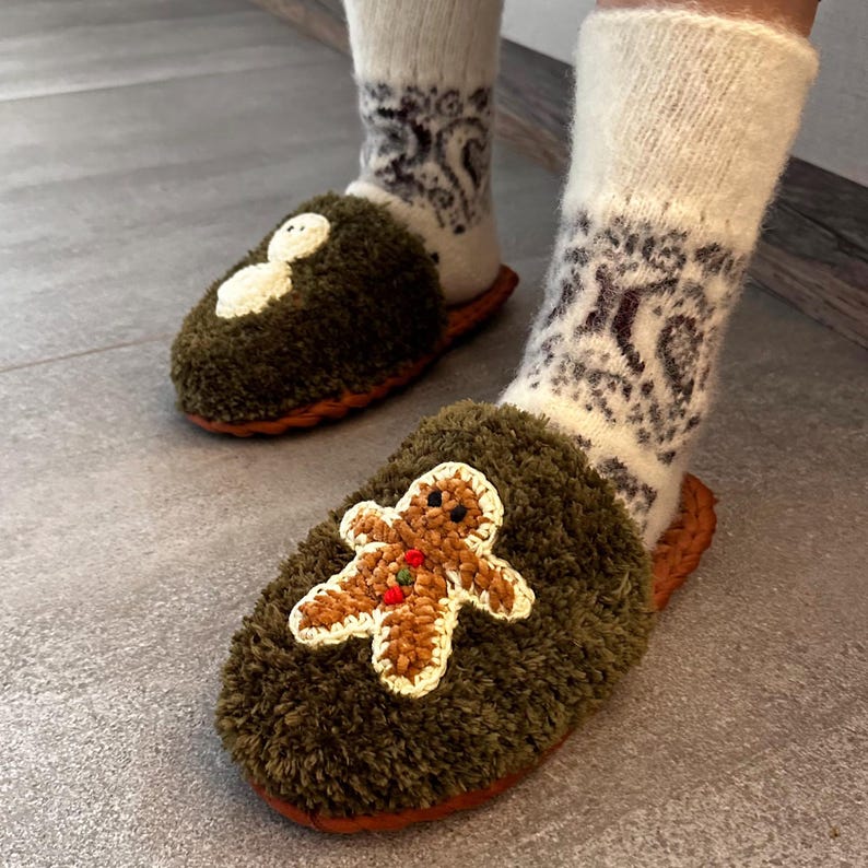 May include: A pair of green fuzzy slippers with a snowman and gingerbread man design. The slippers are worn by a person wearing white socks with a brown pattern.