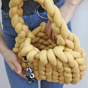 K&ouml;nnte beinhalten: Eine gelbe Handtasche aus geflochtenem Stoff mit runder Form und geflochtenem Henkel. Die Tasche hat einen kleinen schwarz-wei&szlig;en Anh&auml;nger an der Seite. Die Tasche wird von einer Person gehalten, die eine blaue Jeans tr&auml;gt.