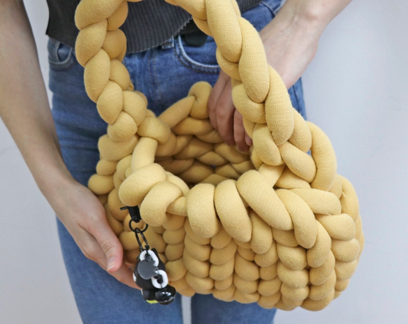 K&ouml;nnte beinhalten: Eine gelbe Handtasche aus geflochtenem Stoff mit runder Form und geflochtenem Henkel. Die Tasche hat einen kleinen schwarz-wei&szlig;en Anh&auml;nger an der Seite. Die Tasche wird von einer Person gehalten, die eine blaue Jeans tr&auml;gt.
