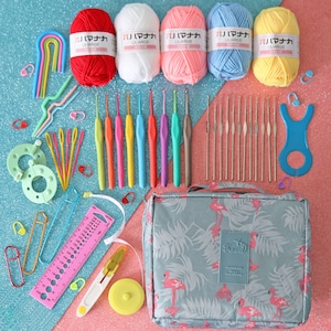 May include: A set of crochet hooks, yarn, and other supplies for crocheting, organised in a grey travel bag with a flamingo print. The yarn is in various colours, including red, white, pink, blue, and yellow. The bag has the word "Travel" printed on it.