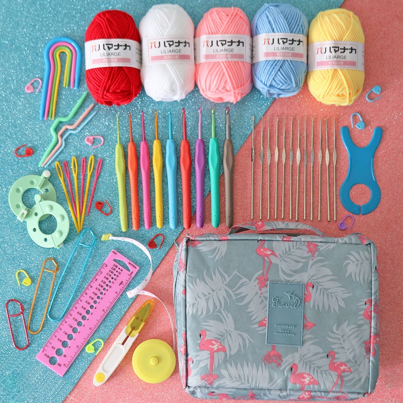 May include: A set of crochet hooks, yarn, and other supplies for crocheting, organised in a grey travel bag with a flamingo print. The yarn is in various colours, including red, white, pink, blue, and yellow. The bag has the word "Travel" printed on it.