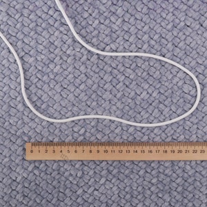 May include: A white elastic cord on a gray textured surface with a wooden ruler marked with numbers 0 to 23.