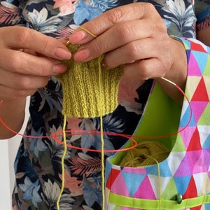 May include: A person is knitting a green knitted item with yellow yarn. The yarn is being held in a colorful bag with a geometric pattern.