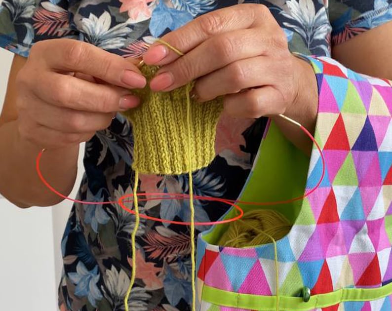 May include: A person is knitting a green knitted item with yellow yarn. The yarn is being held in a colorful bag with a geometric pattern.