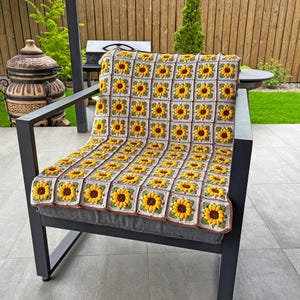 K&ouml;nnte beinhalten: Eine geh&auml;kelte Decke mit Sonnenblumenmuster, drapiert &uuml;ber einem grauen Stuhl. Die Decke zeigt gelbe Sonnenblumen mit braunen Zentren und gr&uuml;nen Bl&auml;ttern auf cremefarbenem Hintergrund. Der Stuhl hat ein dunkelgraues Metallgestell.