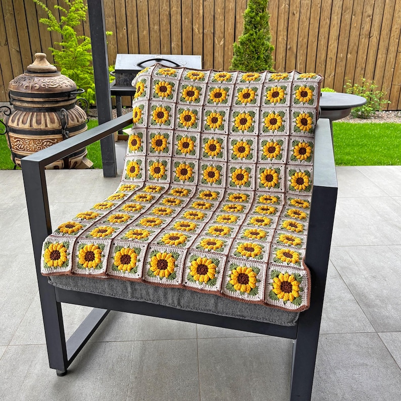K&ouml;nnte beinhalten: Eine geh&auml;kelte Decke mit Sonnenblumenmuster, drapiert &uuml;ber einem grauen Stuhl. Die Decke zeigt gelbe Sonnenblumen mit braunen Zentren und gr&uuml;nen Bl&auml;ttern auf cremefarbenem Hintergrund. Der Stuhl hat ein dunkelgraues Metallgestell.