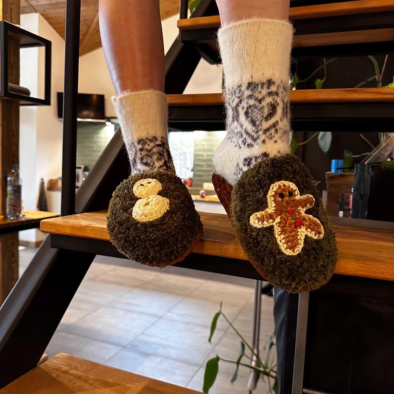 May include: A pair of brown fuzzy slippers with embroidered designs. One slipper features a snowman and the other a gingerbread person.