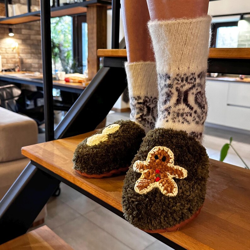 May include: A pair of fuzzy green slippers with gingerbread man appliques. One slipper has a plain gingerbread man applique, while the other has a gingerbread man applique with red and white details. The slippers are worn by a person wearing white socks with a black and white pattern.
