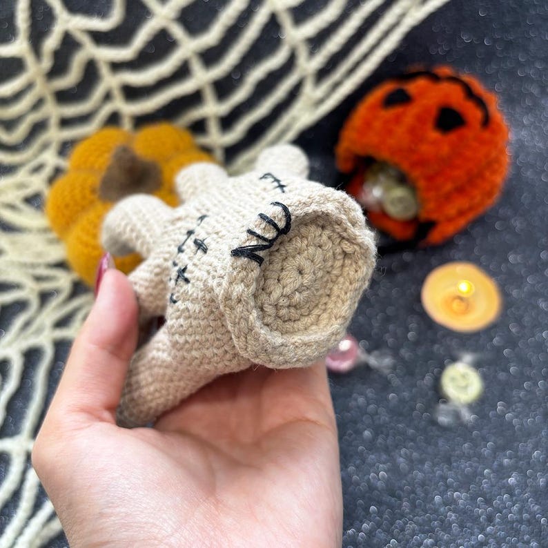 May include: A crocheted Halloween-themed decoration featuring a beige mummy with black stitched details. A small orange pumpkin and a jack-o'-lantern are also visible, suggesting a festive holiday theme. The mummy is held in a hand.