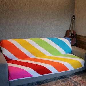 Op de afbeelding: Een kleurrijk gestreept gehaakt deken in tinten roze, rood, oranje, geel, groen, blauw en wit. Het deken is over een grijze bank gelegd.