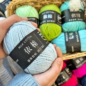 Op de afbeelding: Een close-up van een hand die een lichtblauwe bol garen vasthoudt met het label "Milk Cotton". Andere bollen garen in verschillende kleuren, waaronder groen, blauwgroen en roze, zijn zichtbaar in een gevlochten mand. De bollen hebben zwarte labels met Chinese karakters.
