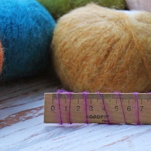 May include: Three balls of yarn in different colors: orange, blue, and a light brown. A wooden ruler with purple thread is in the foreground. The ruler has markings from 0 to 9.