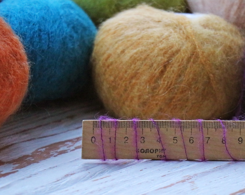 May include: Three balls of yarn in different colors: orange, blue, and a light brown. A wooden ruler with purple thread is in the foreground. The ruler has markings from 0 to 9.