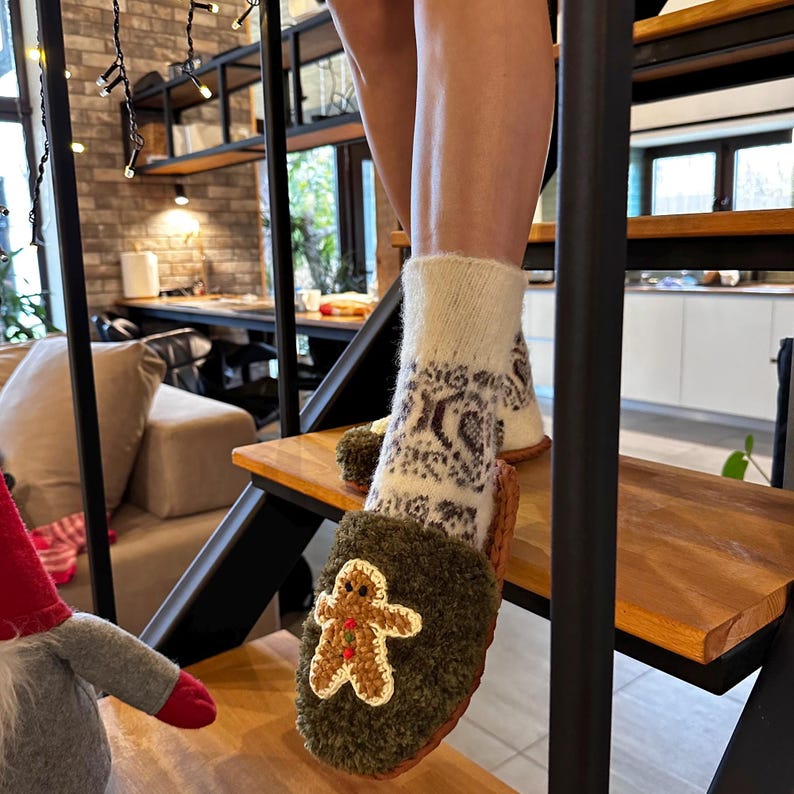 May include: Green fuzzy slippers with a gingerbread man design. The slippers are worn on a person's feet and the person is standing on wooden stairs.