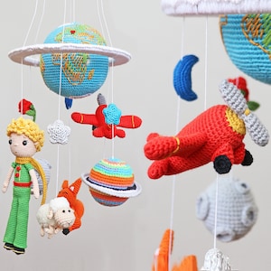 Può includere: Un mobile per neonati all'uncinetto con un personaggio del Piccolo Principe, un aereo rosso, un pianeta blu, un pianeta arcobaleno, una nuvola bianca, una luna grigia, una pecora grigia e una volpe grigia.