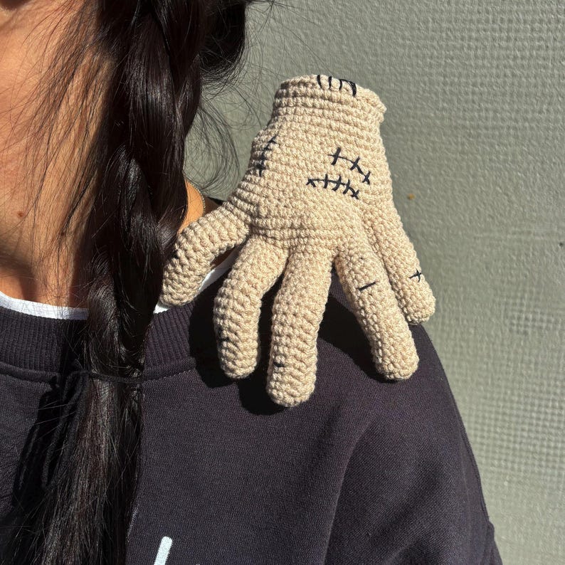 May include: A beige crochet hand with black stitched details. The hand has five fingers and is resting on a dark-colored garment. The stitches create a stylized face on the palm. The hand is a unique, handmade item.
