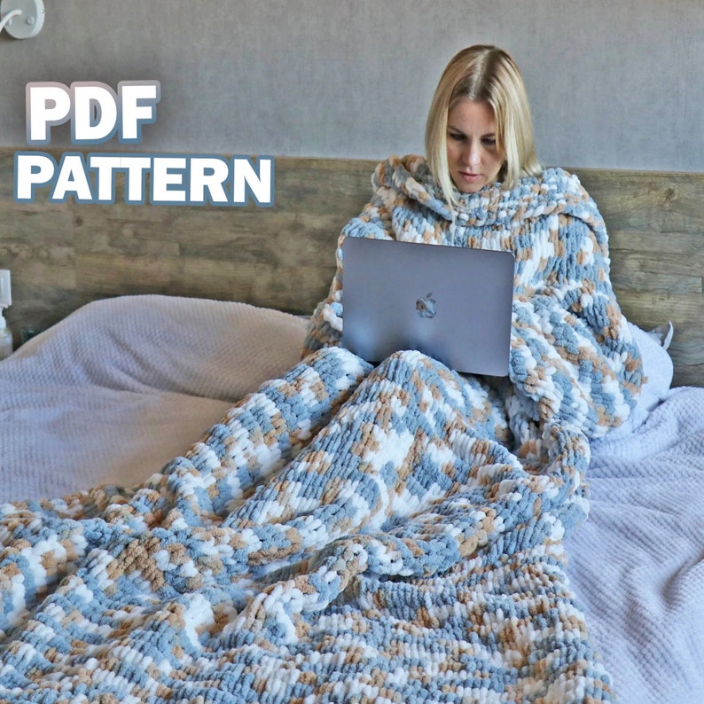 May include: A woman sits on a bed wearing a large, blue, white and brown knitted blanket with a hood. She is using a laptop computer. The text "PDF PATTERN" is displayed at the top of the image.
