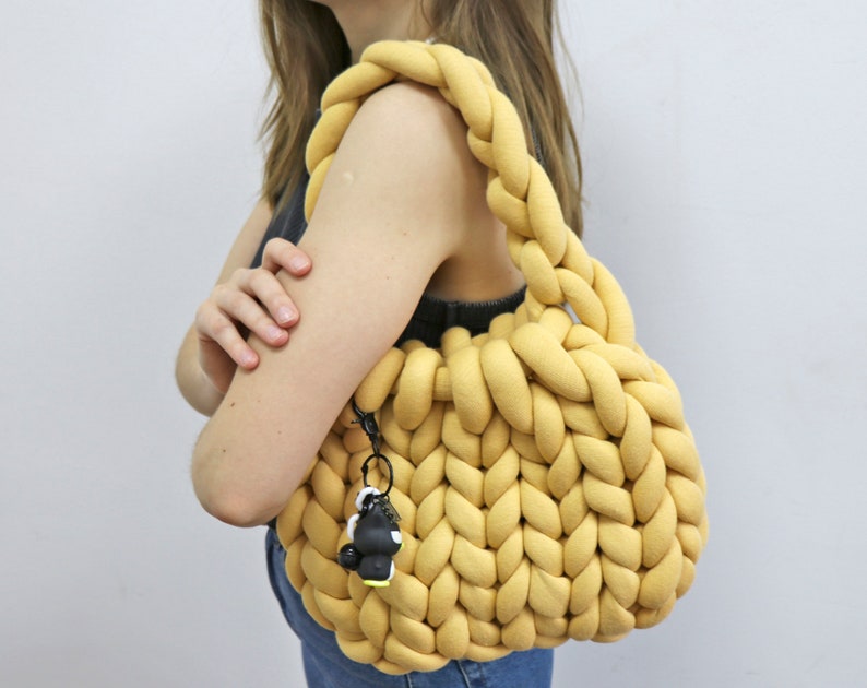 K&ouml;nnte beinhalten: Senfgelbe, grob gestrickte Handtasche mit geflochtenen Henkeln. Die Tasche hat eine runde Form und einen kleinen schwarz-wei&szlig;en Schl&uuml;sselanh&auml;nger an der Seite. Die Tasche ist aus einem weichen, dicken Garn gefertigt.