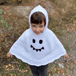 Può includere: Un poncho fantasma bianco all'uncinetto con cappuccio e un volto sorridente in feltro nero. Il poncho è indossato da un bambino. Lo sfondo è una scena sfocata all'aperto con foglie autunnali.