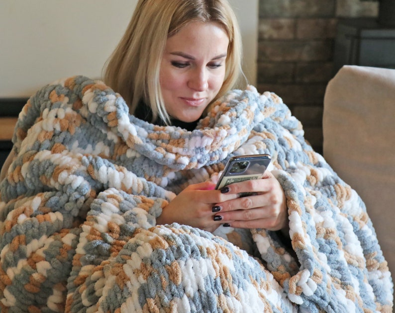 May include: A person wrapped in a chunky knit blanket with a blue, white and beige colour pattern, using a smartphone.