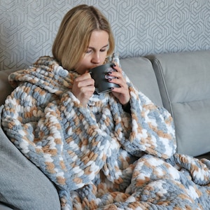 May include: A person wearing a grey, white and brown hooded blanket with a fluffy texture. The person is sitting on a grey sofa and holding a grey mug.