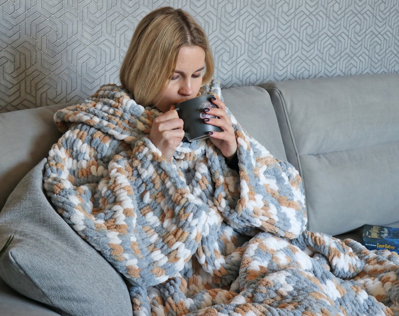 May include: A person wearing a grey, white and brown hooded blanket with a fluffy texture. The person is sitting on a grey sofa and holding a grey mug.