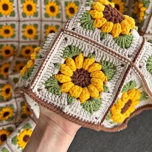K&ouml;nnte beinhalten: Handgefertigte H&auml;keldecke mit einem sich wiederholenden Sonnenblumenmuster. Die Decke besteht aus cremefarbenem, gelbem, braunem und gr&uuml;nem Garn. Die Sonnenblumen sind der Mittelpunkt, mit detaillierten Bl&uuml;tenbl&auml;ttern und Zentren. Die Decke ist quadratisch und wirkt weich und gem&uuml;tlich.