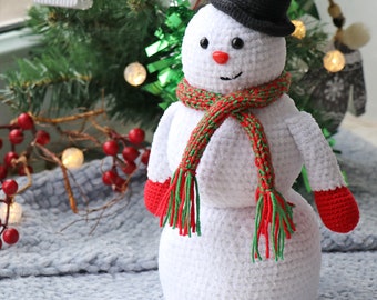 May include: A white crocheted snowman wearing a black hat, a red and green scarf, and red mittens. The snowman is sitting on a gray fuzzy surface.