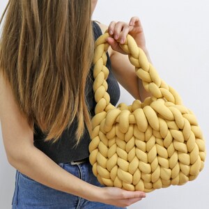 K&ouml;nnte beinhalten: Eine senfgelbe, handgestrickte Tasche mit geflochtenem Henkel. Die Tasche ist aus dickem, weichem Garn gefertigt und hat eine runde Form. Die Tasche wird von einer Person gehalten, die ein schwarzes Tanktop und blaue Jeans tr&auml;gt.