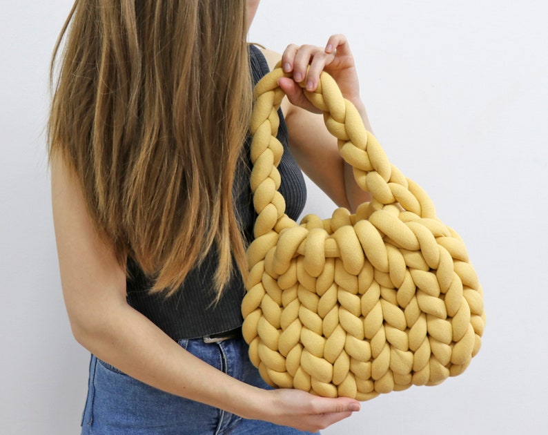 K&ouml;nnte beinhalten: Eine senfgelbe, handgestrickte Tasche mit geflochtenem Henkel. Die Tasche ist aus dickem, weichem Garn gefertigt und hat eine runde Form. Die Tasche wird von einer Person gehalten, die ein schwarzes Tanktop und blaue Jeans tr&auml;gt.