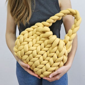 K&ouml;nnte beinhalten: Eine senfgelbe, grob gestrickte Handtasche mit geflochtenem Henkel. Die Tasche ist aus dickem, weichem Material gefertigt und hat eine abgerundete Form. Die Tasche wird von einer Person gehalten.