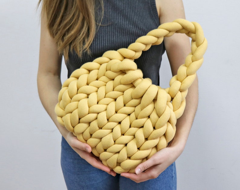 K&ouml;nnte beinhalten: Eine senfgelbe, grob gestrickte Handtasche mit geflochtenem Henkel. Die Tasche ist aus dickem, weichem Material gefertigt und hat eine abgerundete Form. Die Tasche wird von einer Person gehalten.