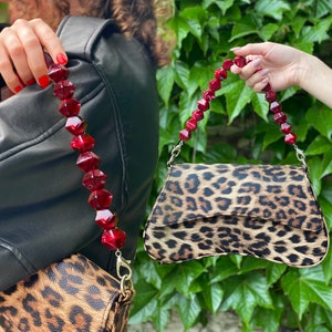 Könnte beinhalten: Eine kleine Handtasche mit Leopardenmuster und einem roten Perlenriemen. Der Riemen besteht aus großen, facettierten Perlen.