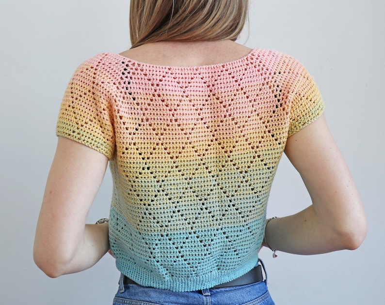 May include: A short-sleeved, crochet top with a scoop neckline. The top features a gradient color scheme, transitioning from pink to yellow to blue. The crochet pattern includes a decorative openwork design.