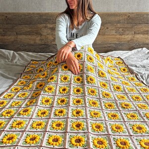 May include: A handmade crochet blanket featuring a repeating sunflower pattern. The blanket has a white background with yellow sunflowers, brown centers, and green leaves. The border is brown.